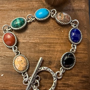 May include: Silver bracelet with oval gemstones in turquoise, green, red, and black. The stones are set in a silver rope-style border and linked by silver. The bracelet has a toggle clasp. The bracelet is suitable for a wrist size of approximately 18 cm.