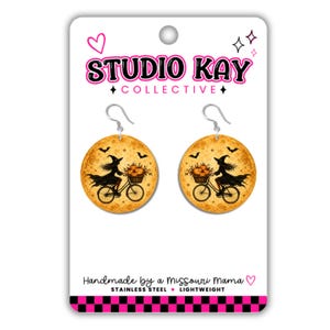 May include: Round dangle earrings featuring a witch riding a bicycle with a basket of pumpkins against a yellow moon background. The display card reads STUDIO KAY COLLECTIVE, Handmade by a Missouri Mama, STAINLESS STEEL, and LIGHTWEIGHT.