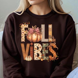 May include: A dark brown sweatshirt with the words "FALL VIBES" in a patchwork design. The letter "O" is replaced with a pumpkin. The sweatshirt is a fall-themed item.