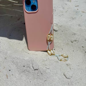 May include: A pink phone case with a blue camera module and a pearl beaded phone strap. The strap features off-white pearls and gold-colored beads strung on a light blue cord. The phone rests on white sand.