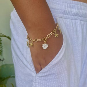 Op de afbeelding: Gouden bedelarmband met bedels in de vorm van zeesterren en schelpen. De armband heeft een schakelketting ontwerp en wordt om de pols gedragen. De bedels geven de sieraden een nautisch thema.