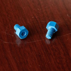 May include: Two small, bright blue bolts with hexagonal heads and threaded shafts rest on a dark wood surface. The bolts are close together, with their heads facing different directions. The bolts are likely used in electronics or small machinery.