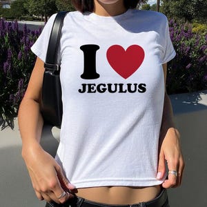 May include: A white short-sleeved t-shirt featuring a graphic print on the chest. The design includes a black letter I, a red heart symbol, and the word JEGULUS in black capital letters. The shirt has a crew neckline and a standard fit.