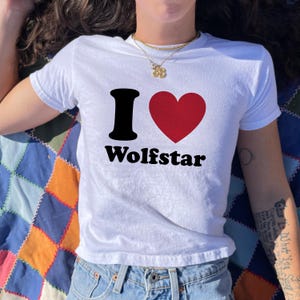 May include: A white short-sleeved t-shirt featuring a black letter I, a red heart symbol, and the black text Wolfstar printed on the front. The shirt is paired with blue denim jeans and a gold-toned chain necklace with a small pendant.