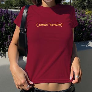 May include: A short-sleeved, crew-neck t-shirt in a deep maroon colour. The centre of the chest features the text (james' version) printed in a bold, golden-yellow font. The shirt is worn with a black shoulder bag featuring a thin strap.