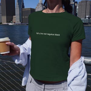 May include: A dark green short-sleeved t-shirt featuring the white text "lol u're not regulus black" printed across the chest. The shirt has a crew neckline and a standard fit, worn with a light blue and white striped button-down shirt layered over it.