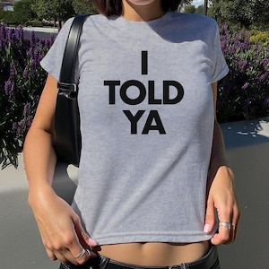 May include: A light grey short-sleeved t-shirt featuring the black text I TOLD YA printed in a bold, sans-serif font across the chest. The garment has a crew neckline and a fitted silhouette, paired with a black shoulder bag with a strap.