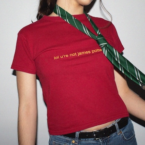 Puede incluir: Una camiseta roja de manga corta y cuello redondo con el texto amarillo "lol u're not james potter" impreso en el pecho. Una corbata verde con rayas plateadas diagonales cuelga alrededor del cuello sobre la camiseta, junto con vaqueros azules.