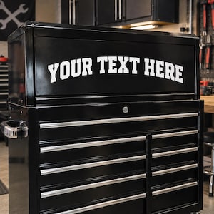 Puede incluir: Un cofre de herramientas de metal negro con un compartimento superior grande que presenta el texto blanco YOUR TEXT HERE impreso en una fuente negrita y arqueada. La unidad incluye múltiples cajones horizontales con tiradores plateados.