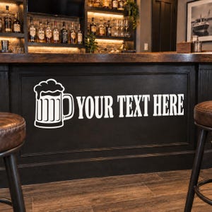 May include: A black bar front featuring a white graphic of a beer mug with foam and the white text YOUR TEXT HERE. The bar has a dark wood top and is flanked by two brown leather stools, with a back bar displaying various glass liquor bottles.