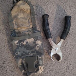 May include: A pair of metal pliers with black handles and a camouflage pouch. The pliers have a curved cutting edge and are made of shiny metal. The pouch is in a digital camouflage pattern.