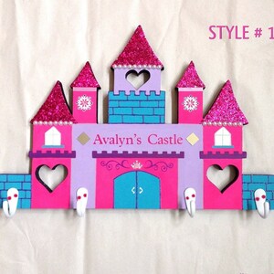 K&ouml;nnte beinhalten: Ein rosa, lila und blauer, aus Holz gefertigter Kleiderhaken in Form eines Schlosses mit einem wei&szlig;en Herz-Ausschnitt und den Worten "Avalyn's Castle" auf der Vorderseite. Der Kleiderhaken hat f&uuml;nf Haken und misst 17 Zoll mal 12 Zoll.