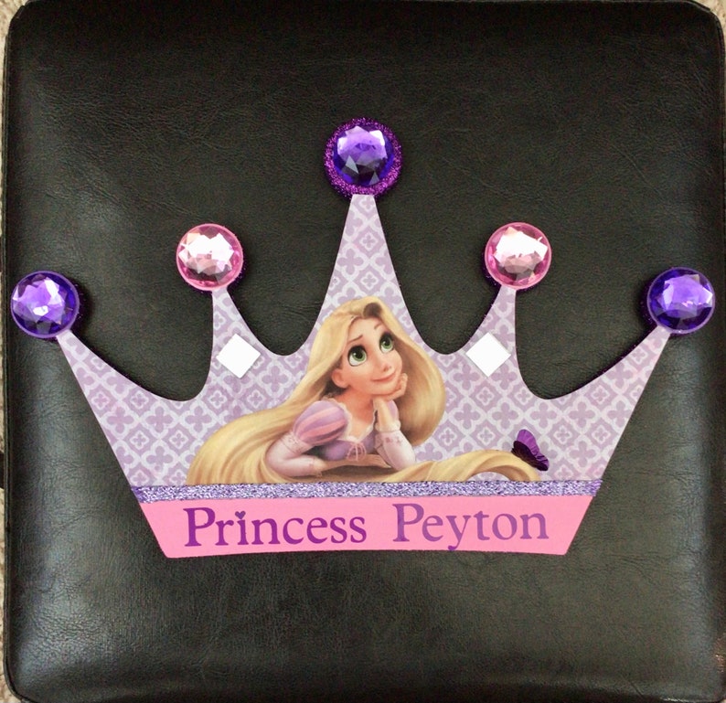 K&ouml;nnte beinhalten: Eine lila und rosafarbene Krone mit Glitzer-Finish und Strasssteinen. Die Krone zeigt ein Bild von Rapunzel aus dem Disney-Film Rapunzel - Neu verf&ouml;hnt. Die Krone tr&auml;gt den Text "Princess Peyton" auf der Vorderseite.