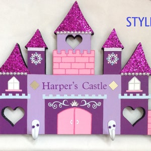 K&ouml;nnte beinhalten: Lilafarbener und rosafarbener, schlosf&ouml;rmiger Schl&uuml;sselhalter aus Holz mit dem Text "Harper's Castle" und vier Haken. Das Schloss hat Glitzerakzente und einen herzf&ouml;rmigen Ausschnitt.