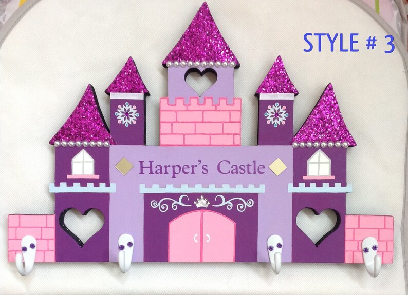 K&ouml;nnte beinhalten: Lilafarbener und rosafarbener, schlosf&ouml;rmiger Schl&uuml;sselhalter aus Holz mit dem Text "Harper's Castle" und vier Haken. Das Schloss hat Glitzerakzente und einen herzf&ouml;rmigen Ausschnitt.