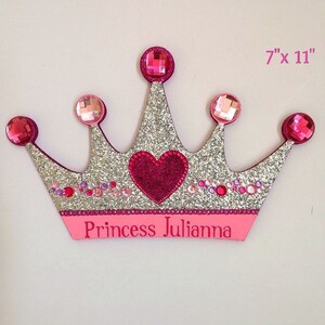 K&ouml;nnte beinhalten: Eine silberne Glitzer-Prinzessinnenkrone mit rosa Akzenten und Strasssteinen. Die Krone hat ein pinkfarbenes Herz in der Mitte und die Worte "Princess Julianna" unten. Die Krone ist 17,8 cm x 27,9 cm gro&szlig;.