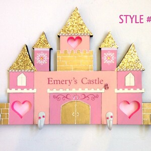 K&ouml;nnte beinhalten: Ein rosafarbener und goldener Holz-Kleiderst&auml;nder in Form eines Schlosses mit dem Namen "Emery's Castle" auf der Vorderseite. Das Schloss hat f&uuml;nf T&uuml;rme, jeder mit einem goldenen Glitzerdach und einem rosa Herz-Ausschnitt. An der Unterseite befinden sich drei Haken zum Aufh&auml;ngen von Gegenst&auml;nden.