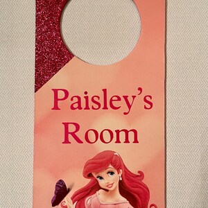 Puede incluir: Un colgador de puerta rosa y rojo con acabado brillante. Presenta una imagen de dibujos animados de Ariel de La Sirenita con una mariposa. El texto "Paisley's Room" est&aacute; impreso en el colgador.