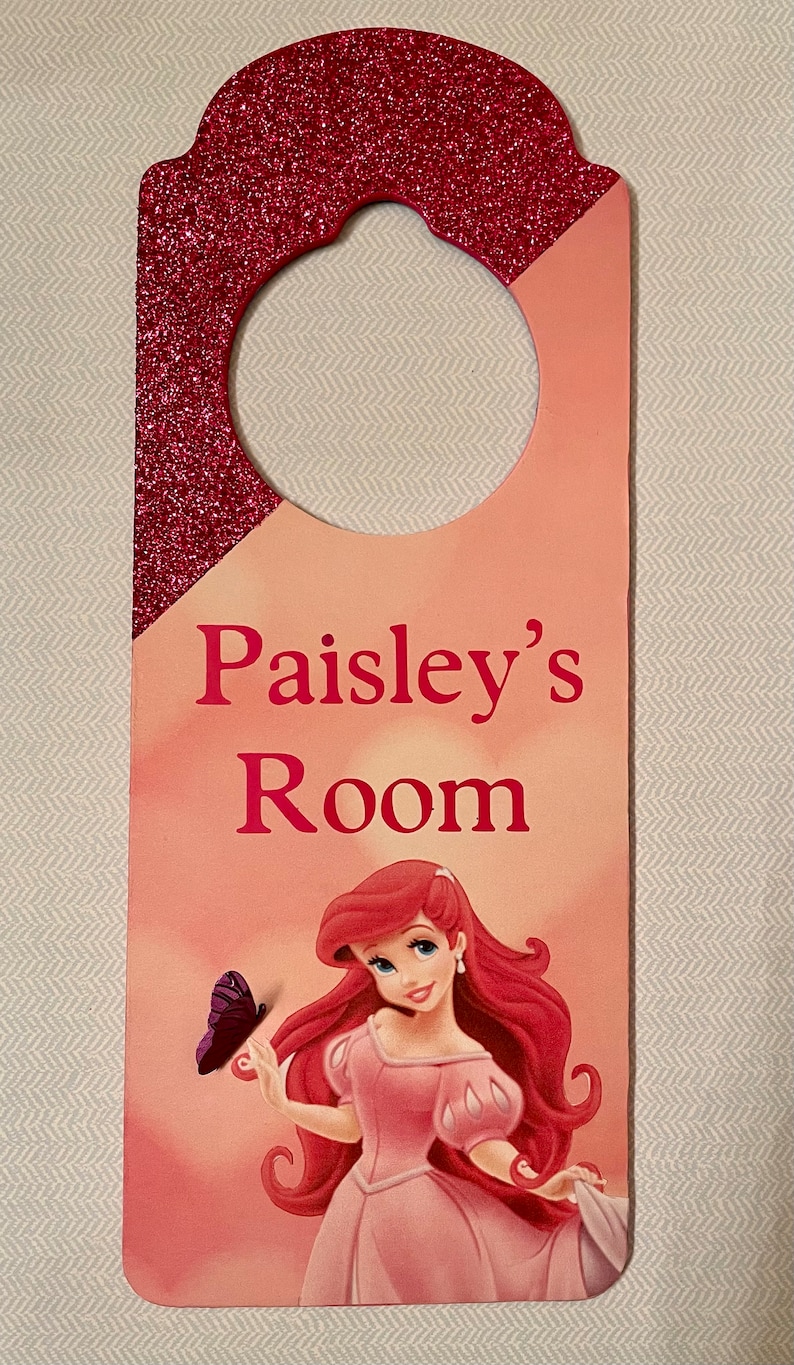 Puede incluir: Un colgador de puerta rosa y rojo con acabado brillante. Presenta una imagen de dibujos animados de Ariel de La Sirenita con una mariposa. El texto "Paisley's Room" est&aacute; impreso en el colgador.