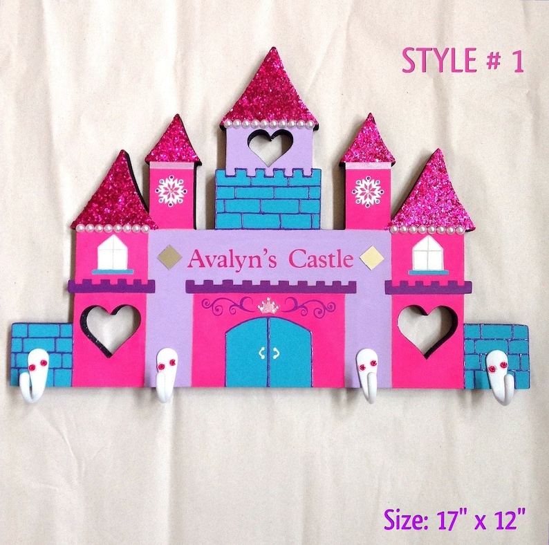 K&ouml;nnte beinhalten: Ein rosa, lila und blauer, aus Holz gefertigter Kleiderhaken in Form eines Schlosses mit einem wei&szlig;en Herz-Ausschnitt und den Worten "Avalyn's Castle" auf der Vorderseite. Der Kleiderhaken hat f&uuml;nf Haken und misst 17 Zoll mal 12 Zoll.