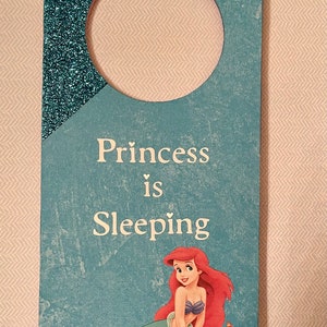 Puede incluir: Un colgador de puerta azul y verde con la parte superior de purpurina. El colgador tiene un agujero para colgar y presenta una sirena con cabello rojo y un cangrejo. El texto en el colgador dice "Princess is Sleeping".