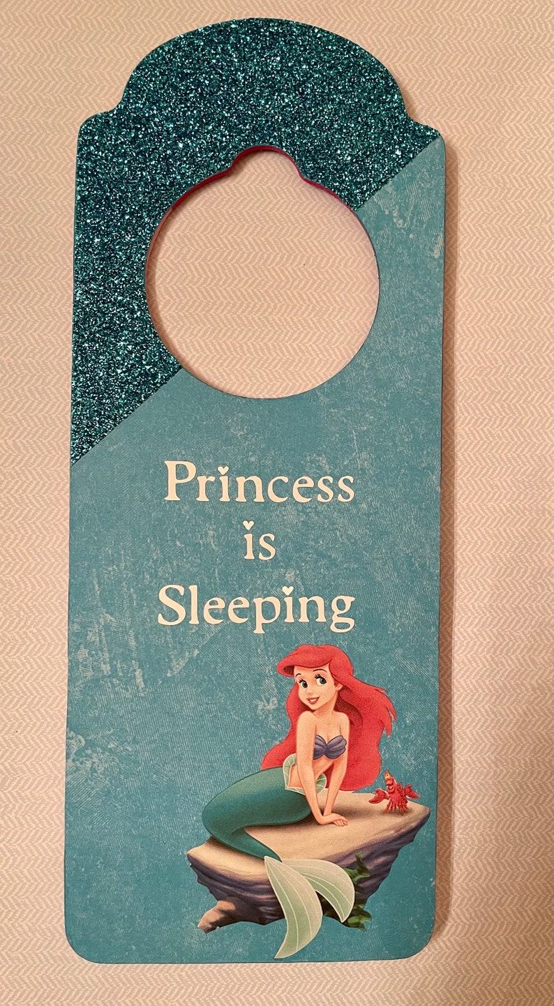 Puede incluir: Un colgador de puerta azul y verde con la parte superior de purpurina. El colgador tiene un agujero para colgar y presenta una sirena con cabello rojo y un cangrejo. El texto en el colgador dice "Princess is Sleeping".