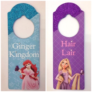 Puede incluir: Dos colgadores de puerta de madera con purpurina y personajes de princesas. El colgador de la izquierda es azul con Ariel de La Sirenita y dice "Ginger Kingdom". El colgador de la derecha es morado con Rapunzel de Enredados y dice "Hair Lair".