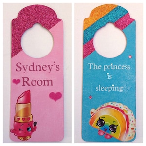 Puede incluir: Dos colgadores de puerta rosa y azul con purpurina. El rosa dice "Sydney's Room" y tiene una caricatura de l&aacute;piz labial. El azul dice "The princess is sleeping" y tiene una caricatura de arco&iacute;ris.