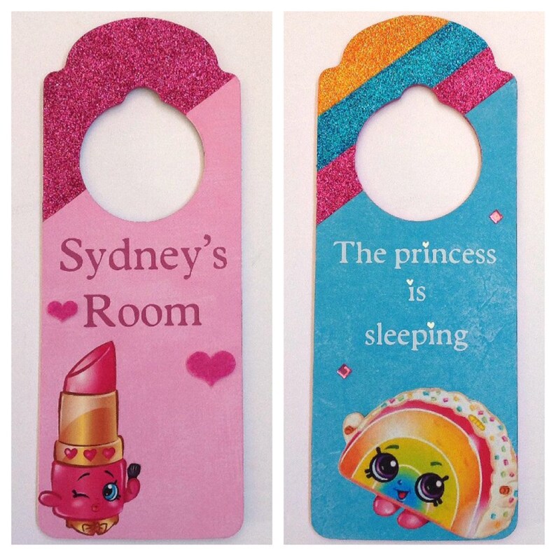 Puede incluir: Dos colgadores de puerta rosa y azul con purpurina. El rosa dice "Sydney's Room" y tiene una caricatura de l&aacute;piz labial. El azul dice "The princess is sleeping" y tiene una caricatura de arco&iacute;ris.