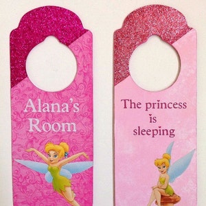 Puede incluir: Dos colgadores de puerta rosas con parte superior de purpurina y dise&ntilde;o de Campanilla. Uno dice "Habitaci&oacute;n de Alana" y el otro dice "La princesa est&aacute; durmiendo".