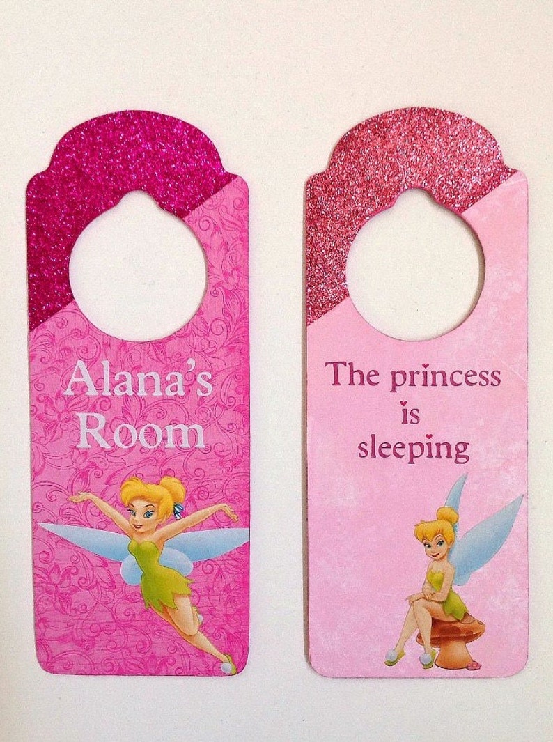 Puede incluir: Dos colgadores de puerta rosas con parte superior de purpurina y dise&ntilde;o de Campanilla. Uno dice "Habitaci&oacute;n de Alana" y el otro dice "La princesa est&aacute; durmiendo".