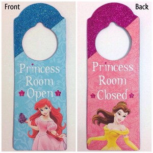 Puede incluir: Un colgador de puerta con tem&aacute;tica de princesa con fondo de purpurina azul y rosa. El frente presenta a Ariel de La Sirenita y el texto "Princess Room Open". La parte posterior presenta a Bella de La Bella y la Bestia y el texto "Princess Room Closed".