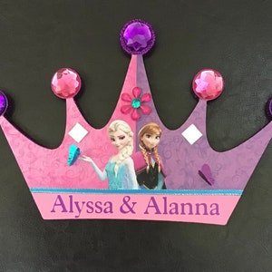 K&ouml;nnte beinhalten: Eine rosa und violette Pappkrone mit einem violetten Glitzerbesatz und Strasssteinen. Die Krone zeigt ein Bild von Elsa und Anna aus dem Film Frozen. Die Krone tr&auml;gt die Namen Alyssa & Alanna auf der Unterseite.