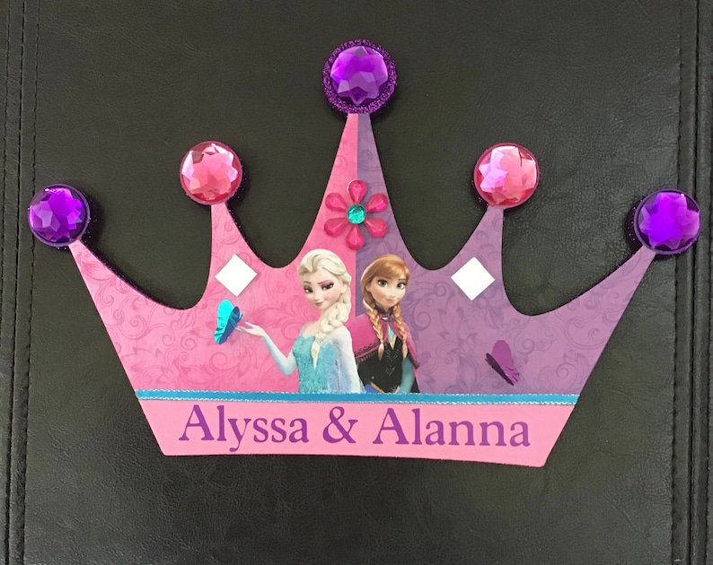 K&ouml;nnte beinhalten: Eine rosa und violette Pappkrone mit einem violetten Glitzerbesatz und Strasssteinen. Die Krone zeigt ein Bild von Elsa und Anna aus dem Film Frozen. Die Krone tr&auml;gt die Namen Alyssa & Alanna auf der Unterseite.