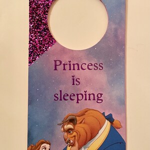Puede incluir: Un colgador de puerta de purpurina morada con un agujero redondo en la parte superior. La mitad inferior del colgador presenta una imagen de dibujos animados de Bella y la Bestia de La Bella y la Bestia de Disney. El texto "Princess is sleeping" est&aacute; impreso en morado en el colgador.