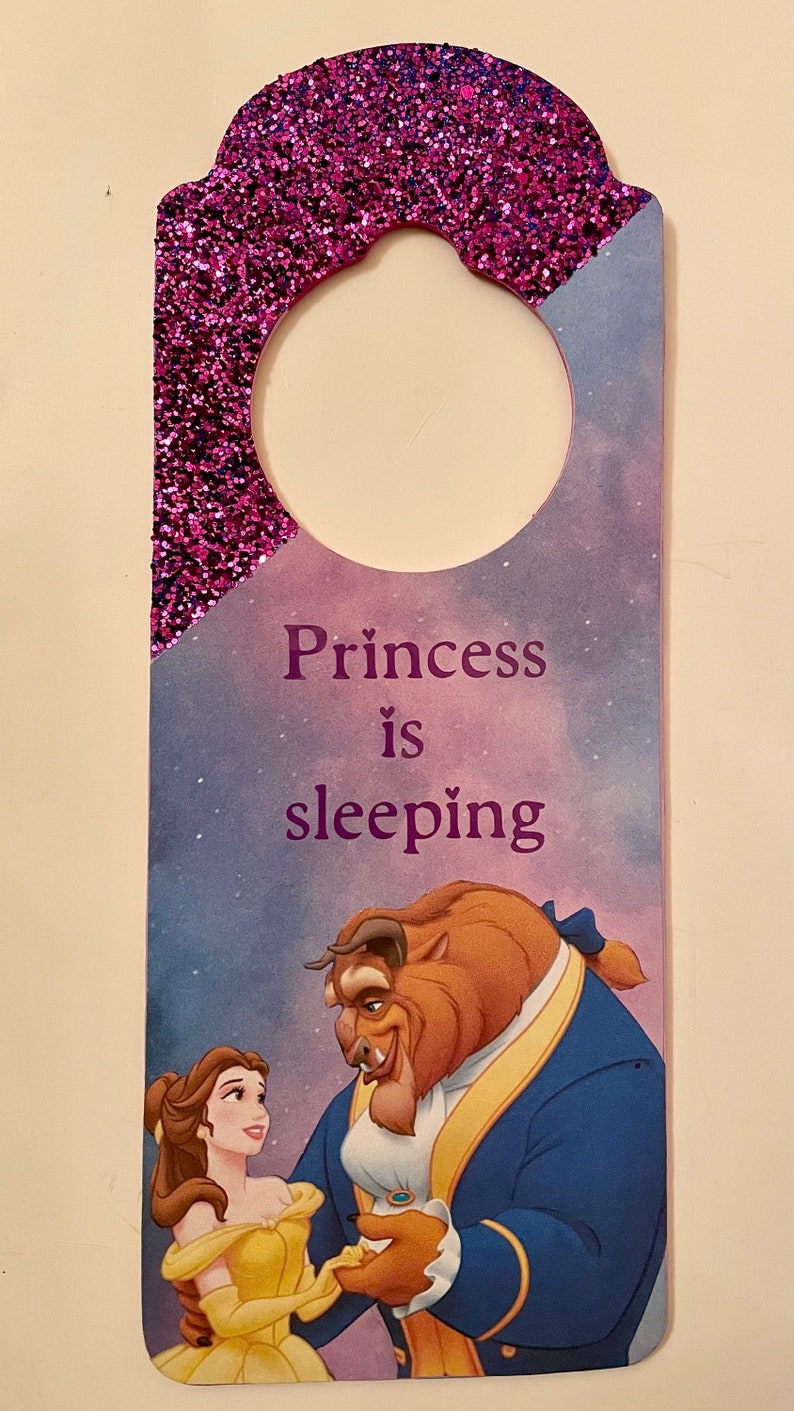 Puede incluir: Un colgador de puerta de purpurina morada con un agujero redondo en la parte superior. La mitad inferior del colgador presenta una imagen de dibujos animados de Bella y la Bestia de La Bella y la Bestia de Disney. El texto "Princess is sleeping" est&aacute; impreso en morado en el colgador.
