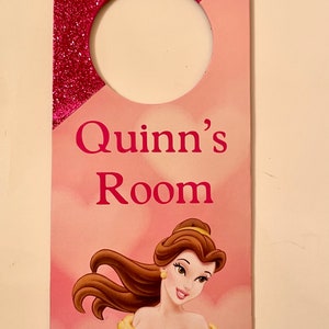 Puede incluir: Colgador de puerta de purpurina rosa y roja con un c&iacute;rculo recortado para colgar. Dice "Quinn's Room" en letras rosas y presenta una imagen de dibujos animados de Bella de La Bella y la Bestia.