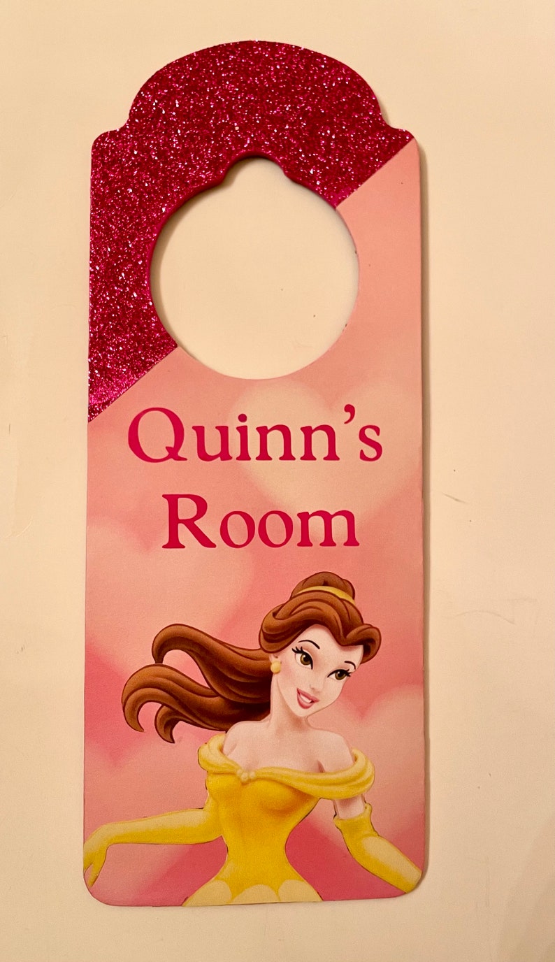 Puede incluir: Colgador de puerta de purpurina rosa y roja con un c&iacute;rculo recortado para colgar. Dice "Quinn's Room" en letras rosas y presenta una imagen de dibujos animados de Bella de La Bella y la Bestia.