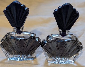May include: Two vintage perfume bottles with black fan-shaped stoppers. The clear glass bottles have a geometric design and are filled with a dark liquid. The bottles are displayed on a light-colored surface.