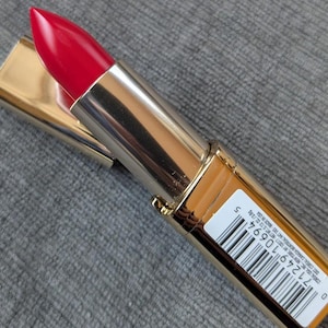 May include: A red lipstick with a gold and silver tube. The lipstick is open, revealing the bright red color. The tube has a barcode and text printed on it. The lipstick is on a gray surface.