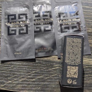 Op de afbeelding: Zwarte huidverzorgingsproductmonsters en een kleine doos. De monsters tonen de tekst "LE SOIN NOIR GIVENCHY" in goud. De doos heeft tekst in meerdere talen en een QR-code.