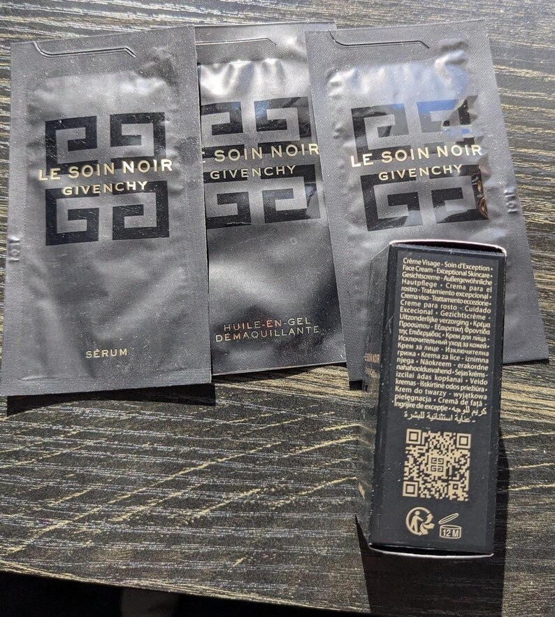 Op de afbeelding: Zwarte huidverzorgingsproductmonsters en een kleine doos. De monsters tonen de tekst "LE SOIN NOIR GIVENCHY" in goud. De doos heeft tekst in meerdere talen en een QR-code.