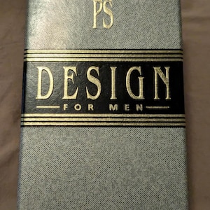 May include: A rectangular cologne box with a textured gray surface. The box features the gold letters "PS" at the top, and "DESIGN FOR MEN" in gold lettering on a black band. The word "COLOGNE" is printed at the bottom.