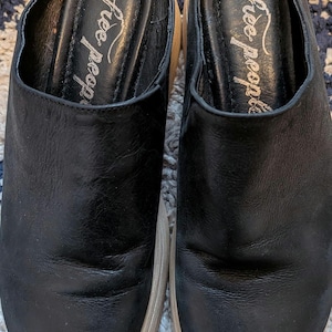 May include: A pair of black leather slip-on mules with a rounded toe and a thick, light-colored platform sole. The interior footbed is black and features the text Free People printed in a white script font on the heel area of each shoe.
