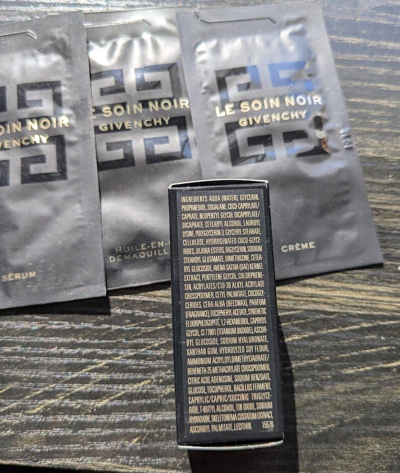 Op de afbeelding: Zwarte zakjes en een kleine zwarte doos met witte tekst. De zakjes zijn gelabeld met "Le Soin Noir Givenchy". De doos bevat een lijst met ingredi&euml;nten. De zakjes zijn voor huidverzorgingsproducten.