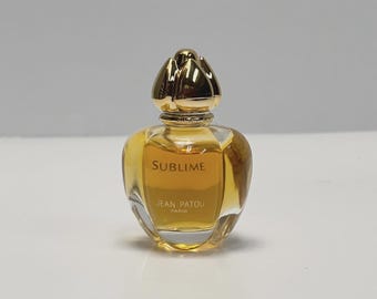 May include: A small, clear perfume bottle with a gold-colored, flower-shaped cap. The bottle contains a golden-yellow liquid and has the word "Sublime" printed on the front, along with "Jean Patou Paris".