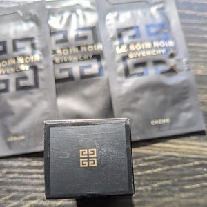 Op de afbeelding: Een zwarte doos met een gouden embleem en verschillende zwarte zakjes. De zakjes zijn voorzien van het opschrift "SOIN NOIR GIVENCHY" in witte tekst. De doos is vierkant en heeft een gouden embleem in het midden.