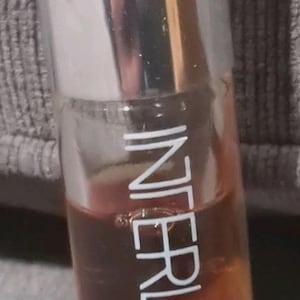 May include: A cylindrical glass perfume bottle with a silver cap, containing an amber-colored liquid. The bottle features white vertical text that reads INTERLUDE and smaller text below that reads SOFT PERFUM, printed directly onto the clear glass surface.