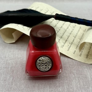 May include: A square glass inkwell with a red base and a dark brown cap, featuring a circular silver emblem with an intricate knotwork design on the front, positioned in front of a black feather quill resting on a piece of aged parchment paper.