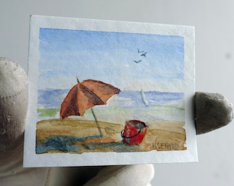 Beach play. Miniature watercolor painting. Original landscape art by artist Ilse Hviid. Blue sky, red bucket and sea, umbrella in the sand.